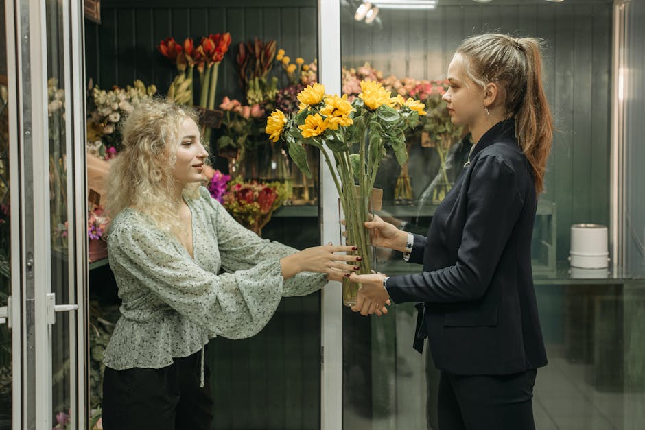 A florist in a dark blazer presenting a tall bouquet of bright yellow flowers, likely sunflowers, with green foliage to a customer inside a flower shop. The customer, a woman with curly blonde hair we