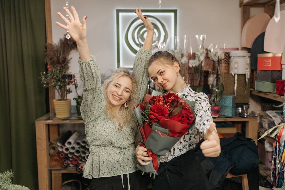 Two women are inside a flower shop, smiling and celebrating. The woman on the right is holding a bouquet of fresh, vibrant red roses wrapped in grey paper with a red ribbon, showing signs of freshness