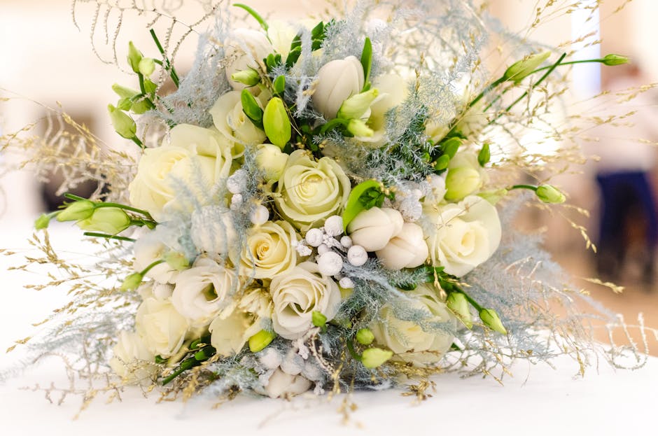 A close-up image of a wedding floral bouquet featuring cream and white roses, surrounded by white tulips, delicate silver-frosted foliage, and various greenery. The bouquet is arranged in a rounded, d