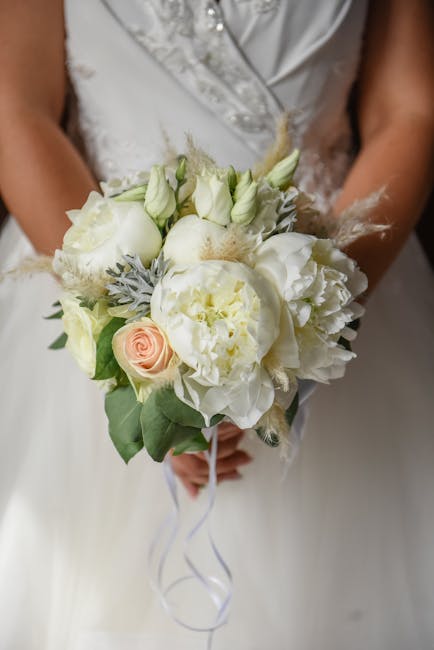 A bride holding a delicate floral bouquet composed of white roses, peonies, and lisianthus, accented with pale green foliage and soft, wispy elements. The bouquet is arranged in a natural, rounded sty