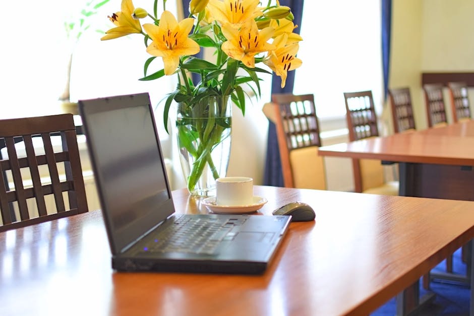 A tall, clear glass vase filled with vibrant yellow lilies featuring large, trumpet-shaped petals and dark stamen details is placed on a polished wooden table. The bouquet is arranged with lush green 