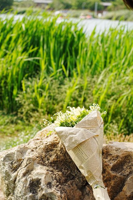 A delicate bouquet of white flowers, possibly freesias or similar blooms, wrapped in crumpled newspaper with visible text, resting on a large textured rock outdoors. The bouquet features fresh, slight