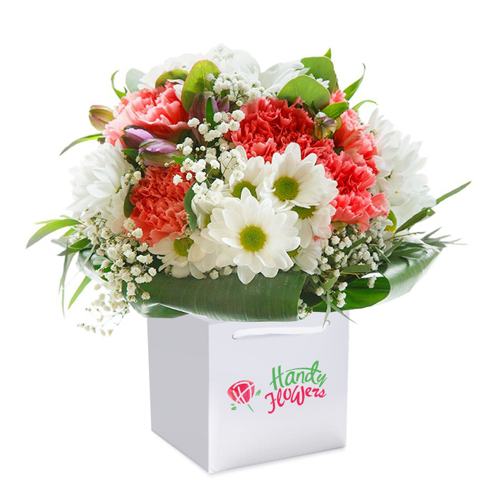Within a flawless white gift bag, this artfully arranged bouquet represents the heart of Wennington's floral tradition-elegant, abundant, and thoughtful. The cluster of soft coral, peach, and bold red carnations stands at the core of the arrangement, their rich ruffled petals adding endless visual interest and a hint of gentle fragrance. Dotted amidst the profusion, snowy white daisies with radiant yellow-green centers catch the light, evoking Wennington's scenic meadows in bloom. Airy white baby's breath flows gently through the bouquet, softening the edges and filling the gaps with light, featherweight accents. Subtle purple-touched buds peek from the left, offering a sense of hidden beauty and organic complexity-a nod to the delicate wildflowers found near the Thames riverside. Around the base, a ring of broad, shiny green foliage creates a polished border, bringing the silhouette together and accentuating the bouquet's lush fullness. Arranged with a florist's careful eye for balance, each element fits harmoniously, resulting in a bouquet that feels joyful, modern, and locally rooted. The sturdy square carry bag boasts practical ribbon handles, making it perfect for a day of gifts around Wennington village or an impromptu celebration by Ferry Lane. This bouquet embodies the expertise and neighbourly spirit of Wennington, thoughtfully prepared to bring delight to every occasion.
