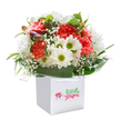 There's a carefree brightness at the heart of this bouquet, as though the light of a Wennington morning has bloomed among the carnations and daisies. Fluffy coral and lively red carnations open wide, each petal edged in fine ruffles that catch the eye and invite the touch. Scattered among the larger blooms, pure white daisies with yellow-green centers create a feeling of joy and openness, their faces lifted like flowers in a sunlit meadow near Wennington Common. Baby's breath, fine as lace, drifts in between, its tiny blossoms lending a sense of innocence and gentle movement throughout. On the left, discreet purple-tinged buds appear, hinting at stories and colours still unfolding, adding a delicate touch of mystery. The composition is wrapped in a collar of bold, glossy green leaves that form an elegant base, all arranged within a smart, white gift bag fitted with long ribbon handles-ready for delivery on a birthday, for congratulating a neighbour, or simply to brighten a Wennington kitchen table. Expertly arranged for both balance and exuberance by Flowers Wennington, this bouquet carries all the freshness and optimism of the local village, promising moments of shared happiness wherever it's delivered.